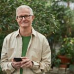 Senior man standing in a greenhouse garden, smiling while using a smartphone. Natural sunlight.