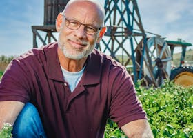 pexels-photo-7457498-7457498 A cheerful bald farmer poses in a lush leafy green field with a vintage windmill in the background.